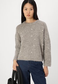 Siv sweater iz dvoslojne povezave z okroglim izrezom, s scattered belimi okrasnimi elementi. Obut je z temno modrimi džinsami, pritrjenimi z črno torbico.