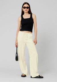 Débardeur noir à côtes, pantalon beige à jambes larges avec cordon de serrage, lunettes de soleil noires et sandales noires. Fond neutre pour le contraste.