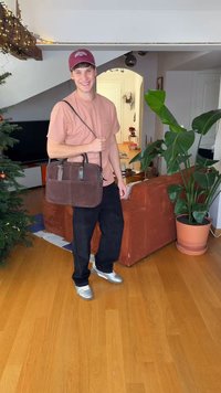 Καφέ δερμάτινη τσάντα χειρός με διπλές λαβές και ιμάντα ώμου, που διαθέτει μπροστινή τσέπη και λεία υφή, κρατιέται από ένα άτομο.