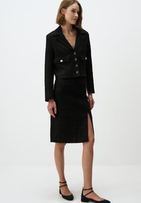 Ensemble noir texturé en deux pièces comprenant une veste courte avec de grands boutons et une jupe longueur genoux avec une fente sur le côté.