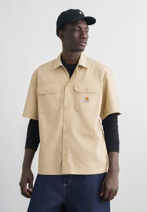 Hombre con camisa beige de manga corta abotonada sobre mangas largas negras, vaqueros azul oscuro y gorra negra, mirando hacia la derecha sobre un fondo liso.