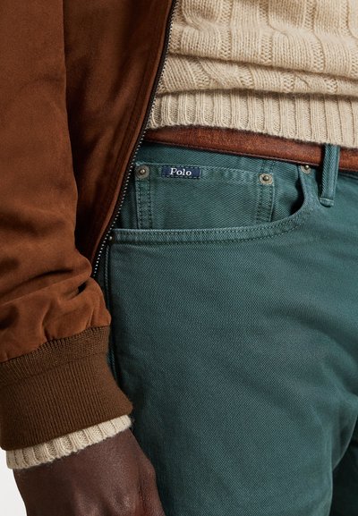 Brūns bumbieru jaka ar ribotām aprocēm, krēmkrāsas adīta džemperis un zaļi Polo bikses ar redzamu etiķeti un metāla pogu akcentiem.