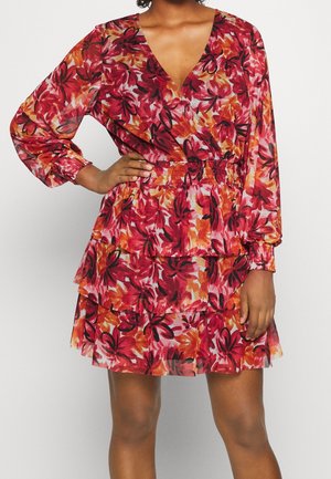 Vrouw draagt een bloemenjurk met lange mouwen en V-hals, met rode, oranje en zwarte patronen, een gerimpelde taille en gelaagde rokpartijen.