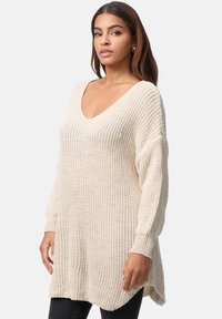 Jersey de punto beige con escote en V, mangas largas y un ajuste relajado. Presenta un punto texturizado y aberturas laterales para facilitar el movimiento.