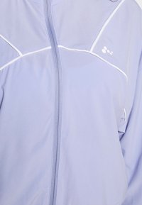 Vaaleanvioletti vetoketjullinen takki, joka on valmistettu sileästä kankaasta. Takissa on valkoisia yksityiskohtia ja logo kaula-aukolla. Yksinkertainen muotoilu ja pehmeä pinta.