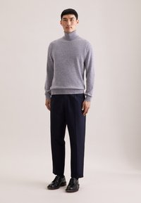 Grauer, weicher, gestrickter Rollkragenpullover kombiniert mit dunkelblauen Hosen und schwarzen Schnürschuhen, vor einem neutralen Hintergrund stehend.