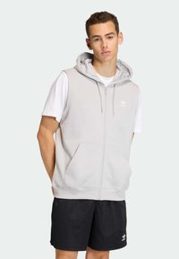 Svetlosivá bezrukávová mikina na zips s prednými vreckami, nastaviteľnou kapucňou so šnúrkou a bielym logom Adidas. V páre s čiernymi šortkami.