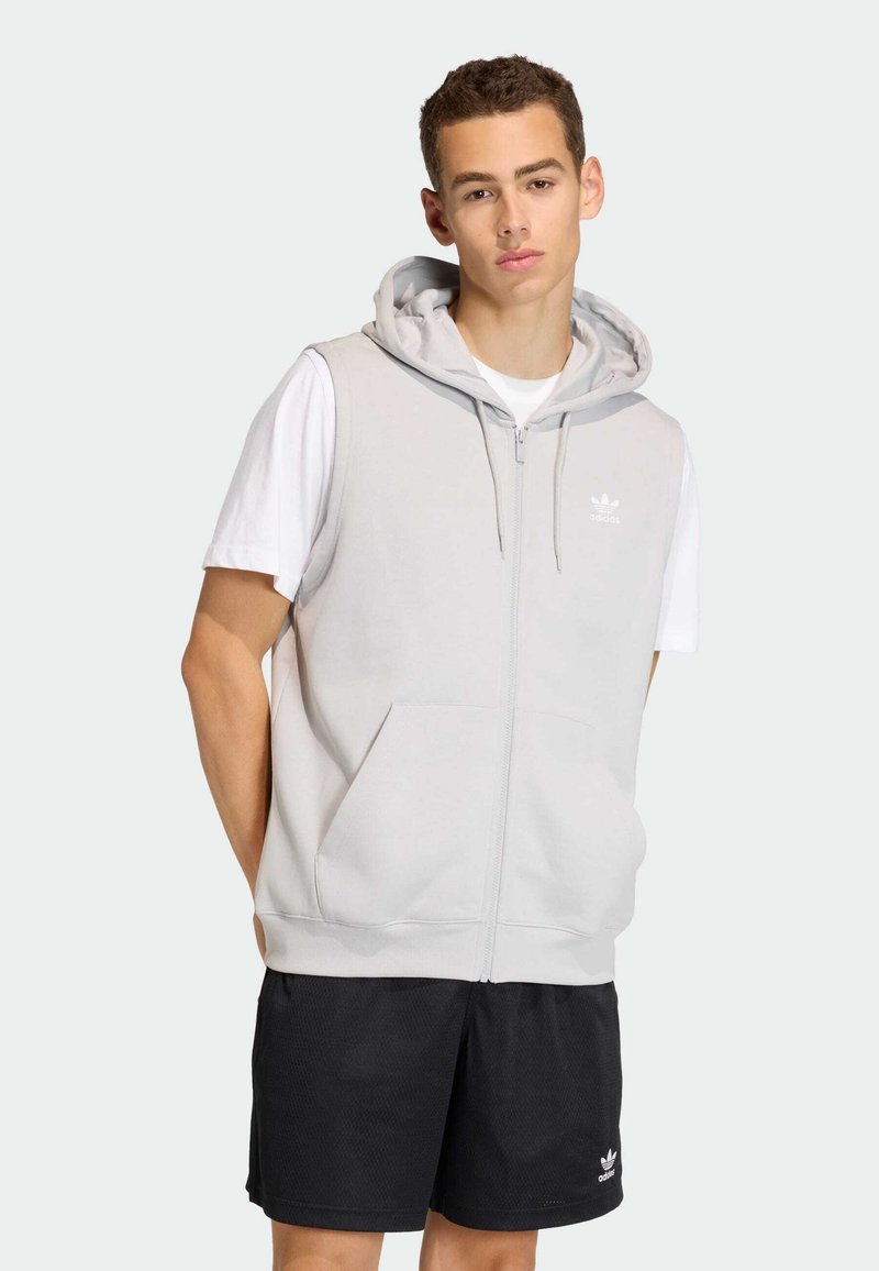 Svetlosivá bezrukávová mikina na zips s prednými vreckami, nastaviteľnou kapucňou so šnúrkou a bielym logom Adidas. V páre s čiernymi šortkami.