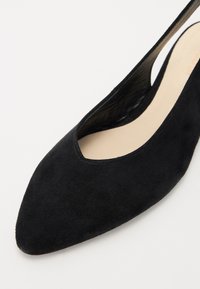 Chaussure à talon haut en suede noir avec un bout pointu et des côtés découpés, dotée d'une doublure intérieure lisse et d'un matériau extérieur texturé.
