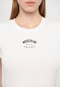 White, ribbed t-shirt featuring a black graphic print that reads "MO5CH1NO 86 23 JEANS" in an arched design on the front.