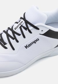 Scarpa da ginnastica bianca realizzata in rete e materiale sintetico, con lacci neri e un'evidente accentuazione nera. Nome del marchio "Kempa" ben visibile.