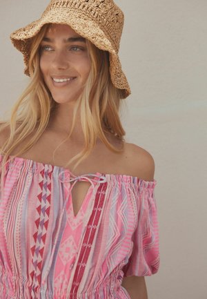 Mladá žena s dlhými blond vlasmi, ktorá nosí tkaný hnedý letný klobúk a ružové šaty s odhalenými ramenami a vzorom, usmievajúca sa a pozerajúca sa do strany.