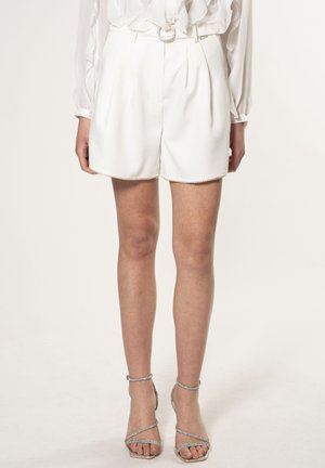 Shorts blancs plissés avec une ceinture présentant une boucle en argent circulaire, associés à des talons aiguilles argentés à lanières. Texture lisse, design arrivant au genou.