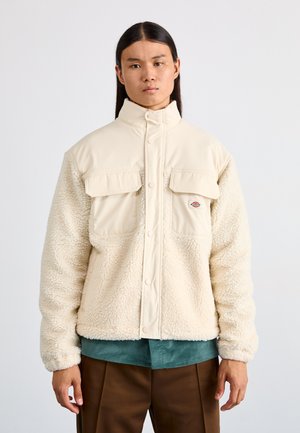 Chaqueta de color crema con una textura suave, que presenta una sección superior firme, botones a presión y dos bolsillos frontales. Diseño suave y cálido.