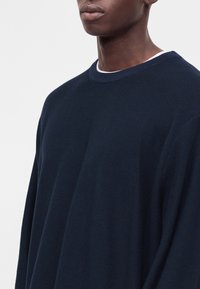 Νεαρό άτομο φοράει σκούνο μπλε υφασμένο πουλόβερ με λευκό εσώρουχο, εμφανίζεται από τους ώμους μέχρι τα χείλη.