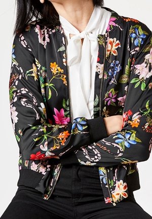 Femme portant une veste bomber noire à fleurs par-dessus un chemisier blanc avec un nœud noué au cou, bras croisés, assise devant un fond uni.