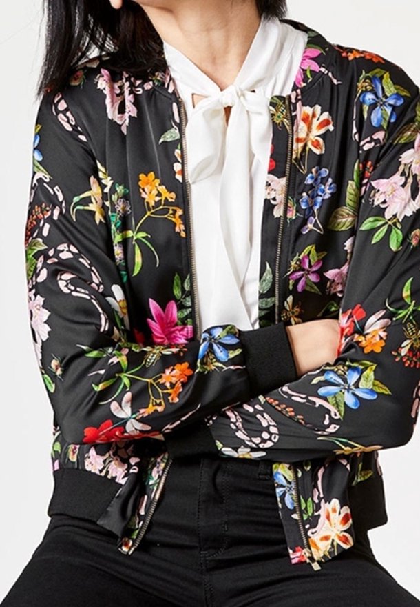 Femme portant une veste bomber noire à fleurs par-dessus un chemisier blanc avec un nœud noué au cou, bras croisés, assise devant un fond uni.