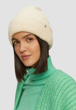 Frau mit braunen Augen und kurzen dunklen Haaren, die eine cremefarbene Strickmütze, einen grünen Rollkragenpullover und eine strukturierte grüne Strickjacke vor einem einfarbigen Hintergrund trägt.