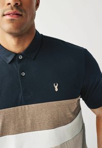 Mornarska polo majica z vodoravnimi črtami v bež in beli barvi, z izvezenim logotipom jelena, dvema gumboma in mehkim bombažnim materialom.