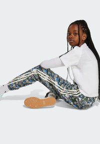 Dieťa v bielom tričku a vzorovaných nohaviciach so zúženým strihom a tromi bielymi pruhmi. Obuv má gumovú podrážku s textúrovaným gripom.