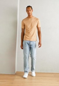 Camiseta ajustada marrón claro, jeans azul claro y zapatillas blancas en una figura de pie. Fondo minimalista, suelo de madera y pared en tonos neutros.