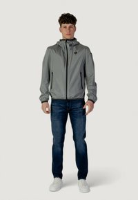 Veste légère grise avec une capuche, fermeture éclair avant, deux poches latérales, associée à un jean bleu foncé et des baskets blanches. Texture lisse, design décontracté.