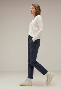 Pull à côtes blanc avec décolleté en V, associé à un pantalon cargo bleu marine équipé de poches latérales. Chaussures : baskets blanches avec semelles beiges.