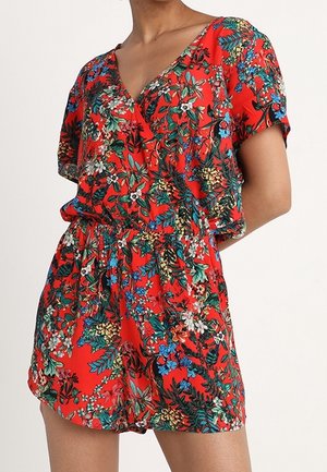 Kvinde iført en rød blomstrede kortærmede romper med V-hals og indsnøret talje mod en ensfarvet baggrund.