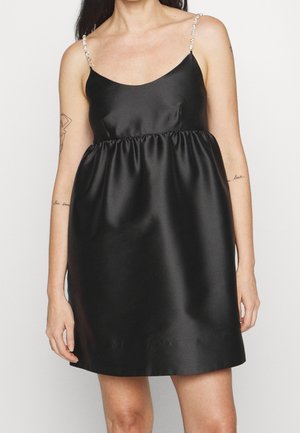 Femme portant une mini-robe noire en satin sans manches avec des fines bretelles perlées, debout devant un fond blanc uni.
