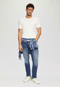 Witte korte mouwen t-shirt, blauw geruit overhemd om de taille geknoopt, blauwe spijkerbroek en witte sneakers. Casual outfit met een ontspannen pasvorm.