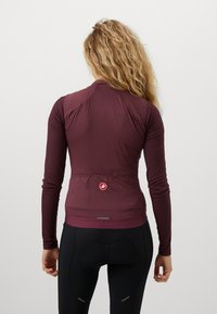 Žena s kovrčavom kosom nosi usku duksericu dugih rukava boje bordo i crne biciklističke kratke hlače s podstavom, okrenuta je leđima.