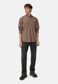 Braun-beige karierte Hemd mit Knöpfen, lange Ärmel, Brusttasche, kombiniert mit dunkelgrauen Jeans und schwarzen Schuhen. Neutraler Hintergrund.
