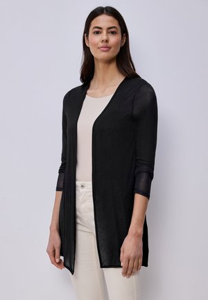 Kobieta o długich ciemnych włosach, ubrana w czarny kardigan z odkrytym przodem, na jasnej bluzce i białych spodniach, stojąca na jednolitym tle.