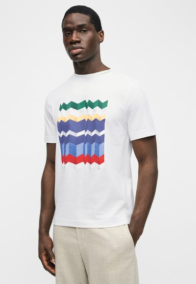 Άνδρας που φοράει λευκό κοντομάνικο μπλουζάκι με πολύχρωμο γεωμετρικό ζιγκ-ζαγκ μοτίβο, συνδυασμένο με ανοιχτό μπεζ παντελόνι, στέκεται μπροστά σε λευκό φόντο.