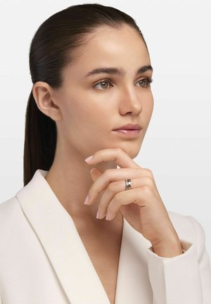 Anello d'argento con gemme trasparenti e nere alternate, indossato su una mano con unghie rosa pallido. Modello in un blazer bianco, sfondo neutro.