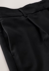 Pantalon noir sur mesure en tissu lisse, avec des plis et des poches latérales. La ceinture comprend une boucle pour la fixation.