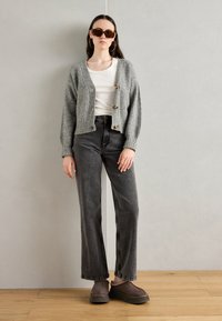 Grå strikket cardigan med tre knapper over en hvit skjorte, kombinert med løse grå jeans og brune plattform sko, aksessorisert med solbriller.