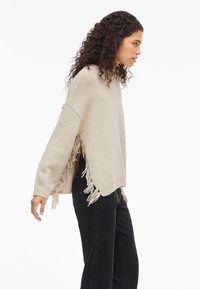 Μπ�εζ πλεκτό πουλόβερ με χαλαρή εφαρμογή, πλαϊνές φούντες, φαρδιές μανίκες και ψηλή λαιμόκοψη, συνδυασμένο με μαύρο παντελόνι. Μαλακή υφή.