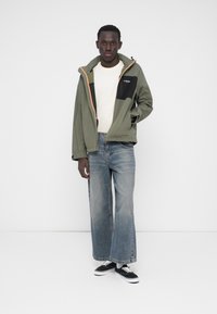 Giacca verde e nera con accenti arancioni, maniche lunghe, tasche con cerniera; abbinata a jeans azzurri a gamba larga e sneakers nere.