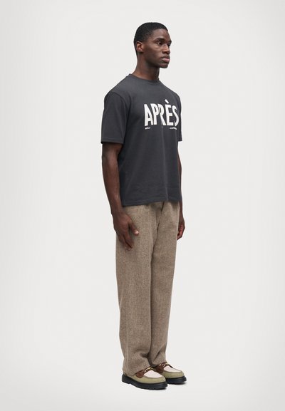 Άνδρας που στέκεται φορώντας μαύρη μπλούζα "APRÈS", μπεζ υφασμάτινο παντελόνι και διχρωμες λουστρίνια με μεταλλική αλυσίδα σε λευκό φόντο.