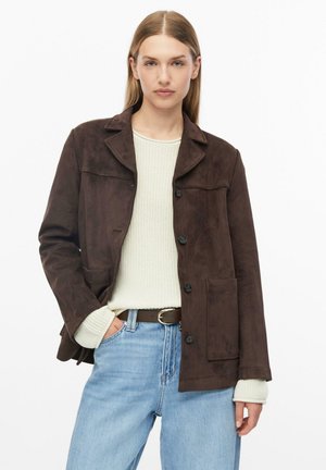 Frau trägt eine braune Wildlederjacke mit Knöpfen, einen cremefarbenen Strickpullover, hellblaue Jeans und einen dunklen Gürtel mit goldener Schnalle.