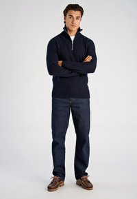 Marineblauer Strickpullover mit Reißverschluss, gerippten Bündchen und Saum, kombiniert mit dunklen Jeans und braunen Bootsschuhen mit Schnürsenkeln und strukturierter Sohle.