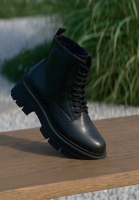 Stivale alla caviglia in pelle nera con lacci frontali, suola in gomma spessa, dettagli testurizzati e una linguetta posteriore per una facile calzata.