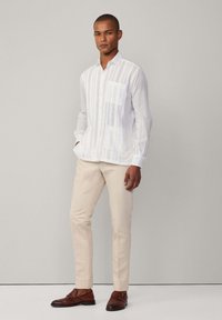 Biała koszula z długim rękawem w pionowe paski, pojedyncza kieszeń na piersi, kołnierzyk zapinany na guziki, zestawiona z jasnobeżowymi spodniami i brązowymi butami.