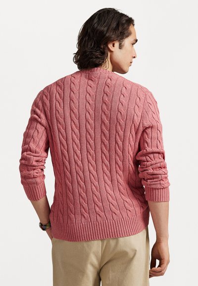 Πλεκτό πουλόβερ σε θαμπό ροζ με ριμπ μανσέτες και στρίφωμα, με υφή σε σχέδιο. Φορεμένο πάνω από μπεζ παντελόνι, προβολή από πίσω.