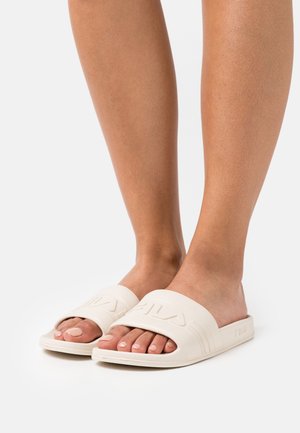 Sandalias de goma blancas de tipo deslizante con una tira ancha que presenta un logo en relieve y ranuras; la textura suave y el diseño de punta abierta destacan el pie.