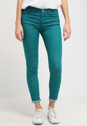 Personne portant un jean skinny bleu canard à revers, des baskets blanches basses et une chemise blanche, debout devant un fond uni.