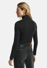 Frau mit blonden Haaren, die ein schwarzes Langarm-Turtleneck und dunkelgraue Jeans mit einem schwarzen Gürtel trägt, von hinten vor einem weißen Hintergrund gezeigt.