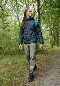 Czarna pikowana kurtka z kapturem, zielone spodnie cargo i czarne trampki. Osoba niesie turkusowy plecak w leśnym otoczeniu.