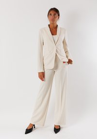 Completo beige su misura con maniche lunghe, revers a punta e tasche frontali, abbinato a pantaloni a gamba larga coordinati e tacchi a punta neri.